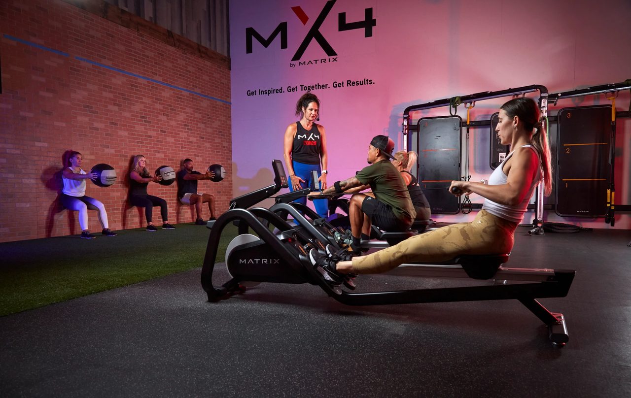 An MX4 coach leading a small-group class on rowers and wall-ball squats